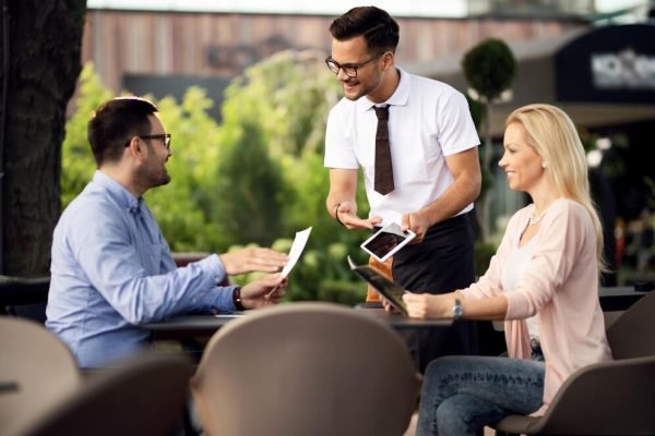 happy-waiter-showing-guests-menu-digital-tablet-while-helping-them-decide-what-order-outdoor-restaurant_637285-545